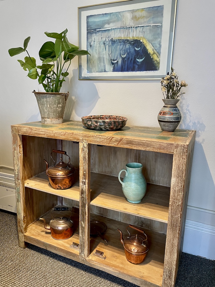 Wooden shelving unit with jugs and plant with a blue painting on the wall